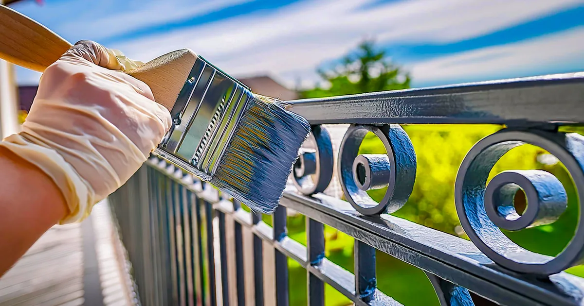 Malarz nakłada pędzlem szarą farbę alkidową na metalową balustradę balkonu