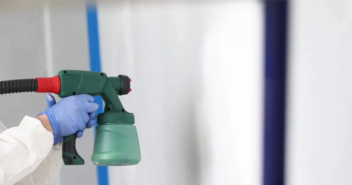 Malarz w kombinezonie i masce natryskuje farbę elektrycznym pistoletem malarskim na ścianę