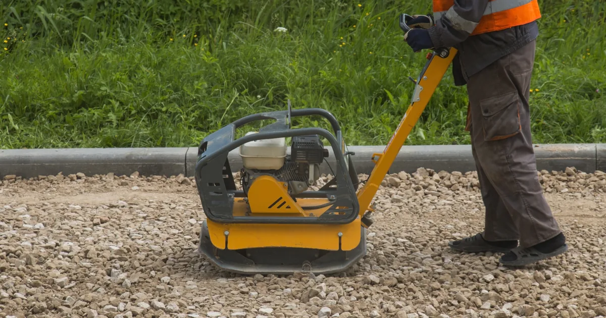 Robotnik zagęszcza warstwę kruszywa zagęszczarką płytową przy krawędzi trawnika
